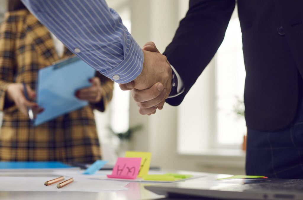 Close-up of two people shaking hands across a desk covered with papers, pens, and sticky notes, with a third person in the background holding a clipboard, indicating a business meeting or job placement agreement.