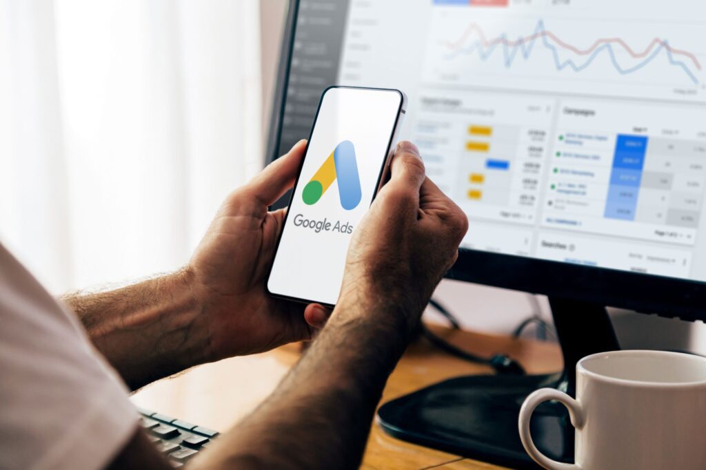 A person holding a smartphone displaying the Google Ads logo, with a desktop screen showing campaign performance data—highlighting HIDM’s training in Google Ads and PPC campaigns, covering ad creation, bidding strategies, performance tracking, and ROI optimization.
