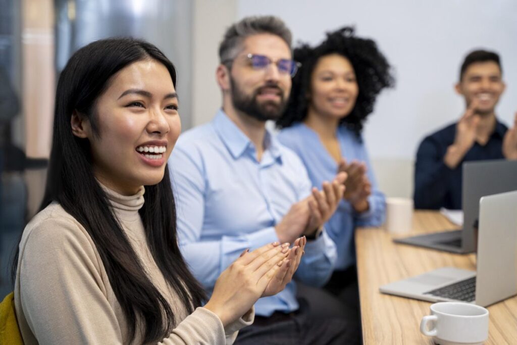 Group of diverse professionals sitting at a conference table, smiling and clapping during a seminar or team meeting. The image reflects positive learning environment, digital marketing workshops, and interactive sessions offered by HIDM – Hisar Institute of Digital Marketing.
