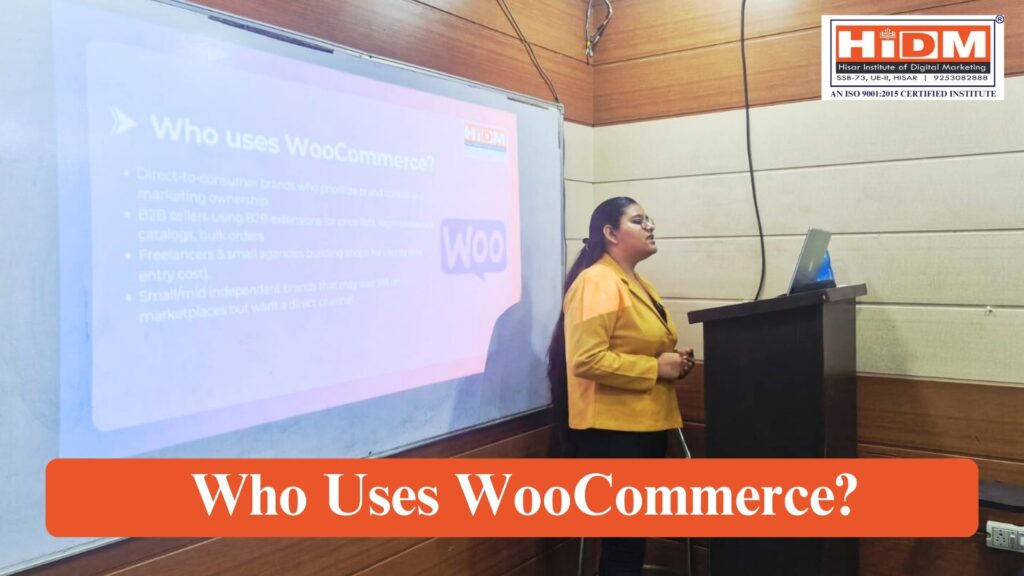 A presenter is delivering a seminar at HiDM (Hisar Institute of Digital Marketing) while explaining a presentation slide titled "Who Uses WooCommerce?" The slide discusses different types of businesses that benefit from WooCommerce, including direct-to-consumer brands, B2B sellers, freelancers, small businesses, and independent brands. A podium with a laptop and projector screen are visible in the classroom.