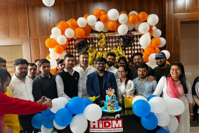 A group of people gathered behind a HiDM themed cake surrounded by orange, white, and blue balloon decorations