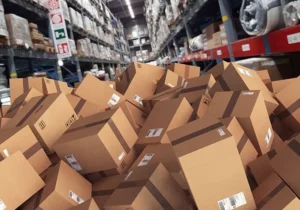 A warehouse aisle filled with a large pile of brown cardboard shipping boxes scattered across the floor, with tall storage racks stocked with inventory in the background, illustrating warehouse overflow, inventory management challenges, or logistics issues.