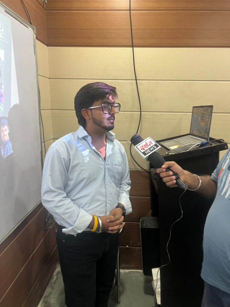Goutam giving a media interview after conducting a seminar on VN AI Editor at HiDM. He is speaking into a news microphone while standing beside the presentation screen and laptop setup in the seminar hall.